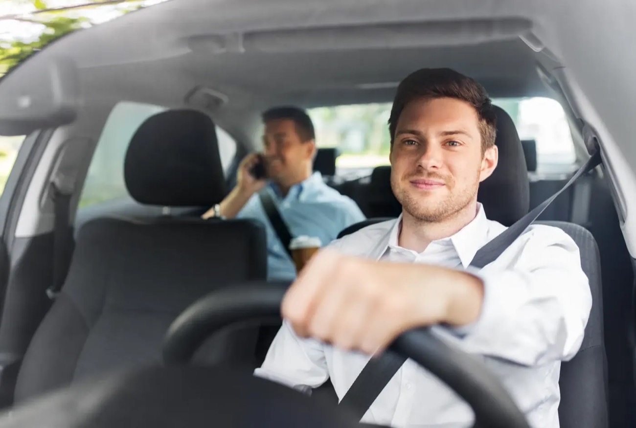 A driver at the wheel of a car wearing a fastened seat belt, with a passenger seated behind, both in daylight.