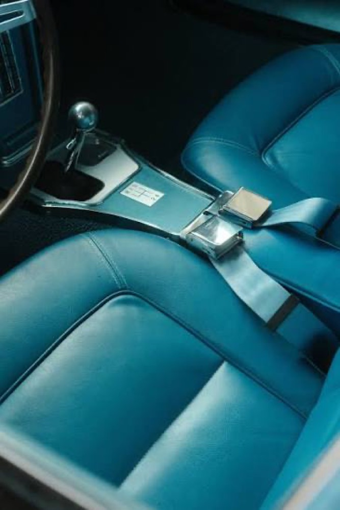 The interior of a classic car with blue leather seats and a chrome lap belt buckle resting on the centre console.