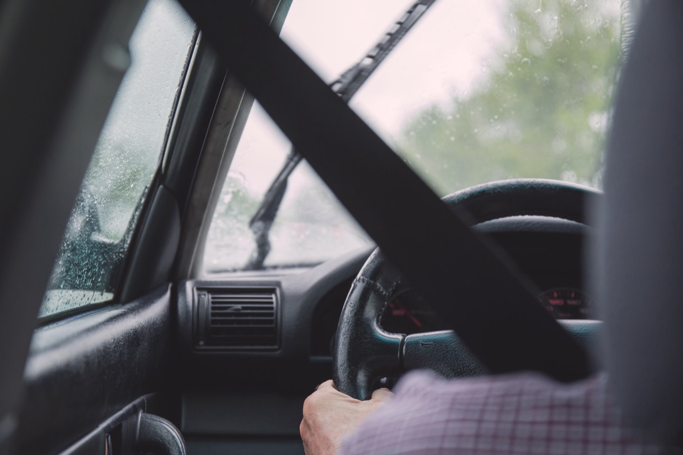 A driver&rsquo;s view from inside a vehicle, hands at the steering wheel with the seat belt running across the shoulder and the road visible through the windscreen.